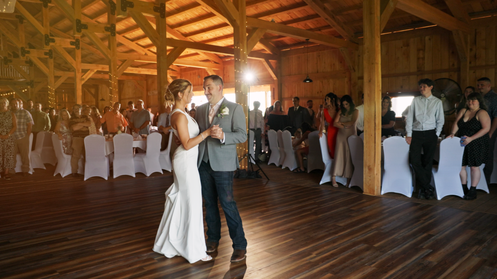 Newly married couple during their first dance. The barn is light with ambient lights, a spotlight, and sun coming from the doors. The guests stand watching in awe as the couple dances. 